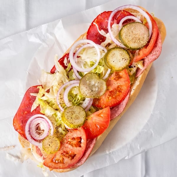 A large deli sub sandwich with salami, lettuce, tomatoes, onions, and pickles on white paper.