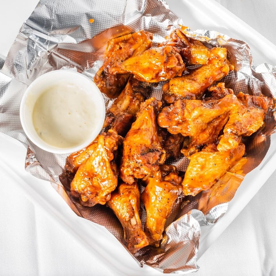 A takeout container filled with saucy chicken wings and a small cup of dipping sauce.