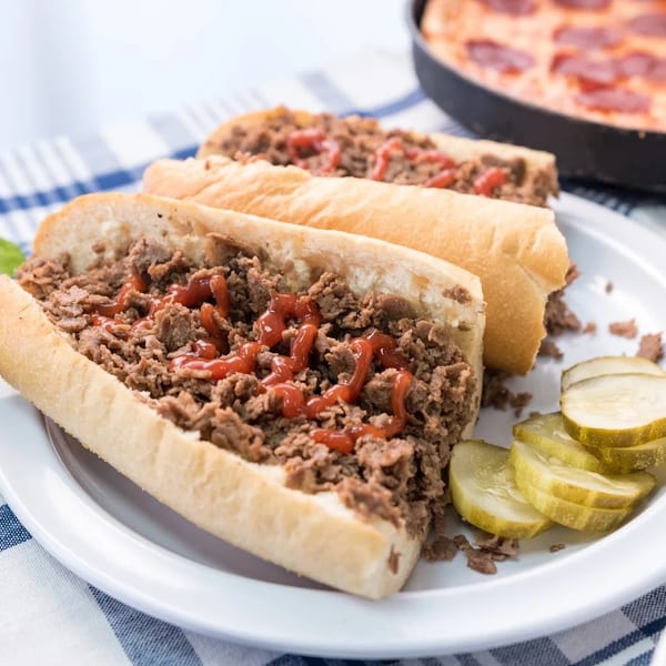 Two cheesesteak sandwiches with ketchup, pickles, a side salad, and a pepperoni pizza.