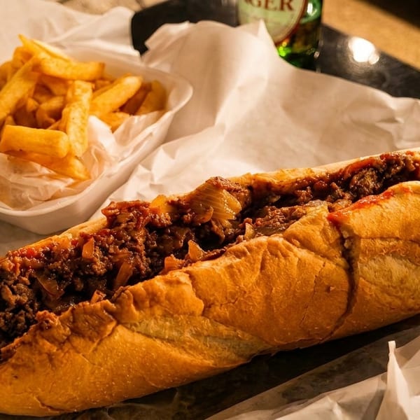 A large cheesesteak sandwich with onions and ketchup, next to a container of golden french fries.