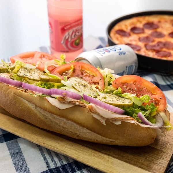 A turkey sub sandwich with lettuce, tomato, pickles, and red onion, next to a pepperoni pizza.