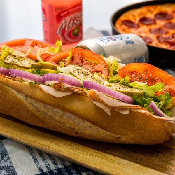 A turkey sub sandwich with lettuce, tomatoes, onions, and pickles, next to a pepperoni pizza and sodas.