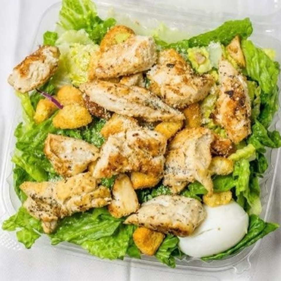 Grilled chicken Caesar salad with croutons and egg in a clear takeout container.